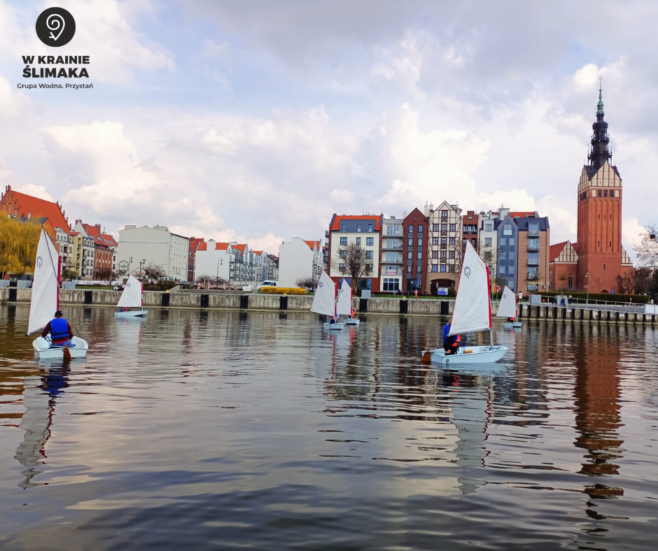 Widok na miasto Elbląg znad tafli wody. Na rzece pływa kilka łodzi z ludźmi. W oddali widać nowoczesne kamienice i kościół.
