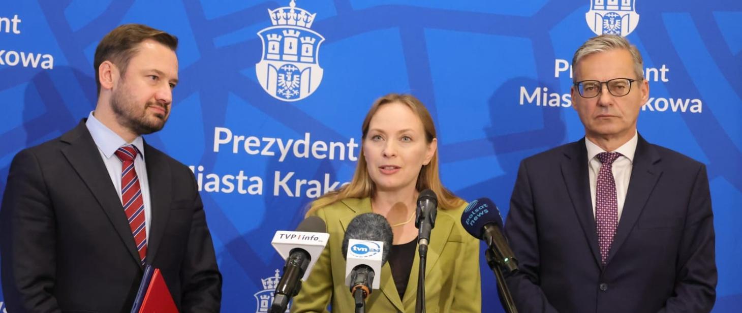 Trzy osoby stoją obok siebie. W środku przed mikrofonami stoi minister Katarzyna Pełczyńska-Nałęcz.