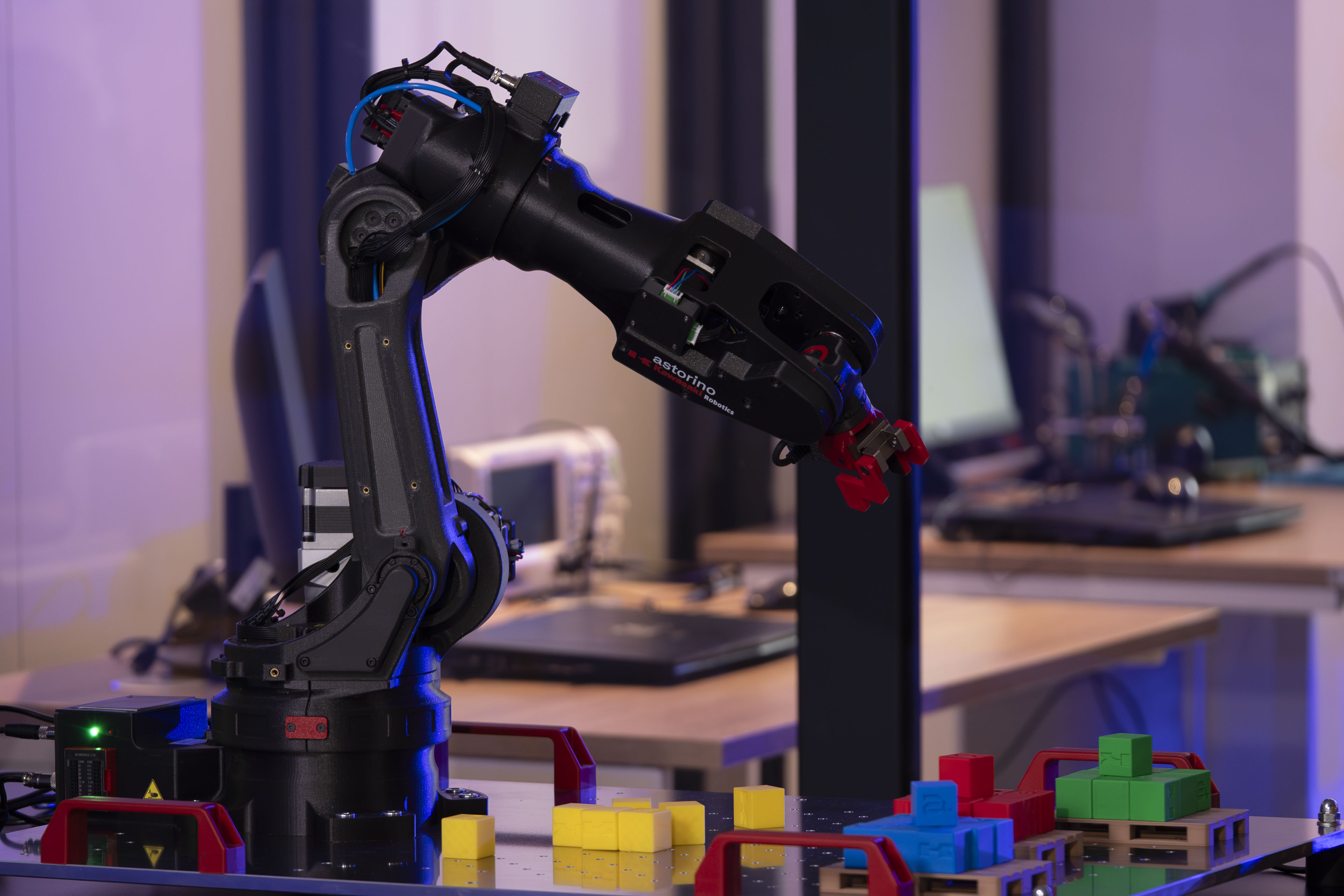 Czarny robotyczny manipulator z chwytakiem pracujący na stole laboratoryjnym, obok którego leżą kolorowe klocki (żółte, czerwone, niebieskie i zielone) ułożone w grupy. W tle widoczne komputery.