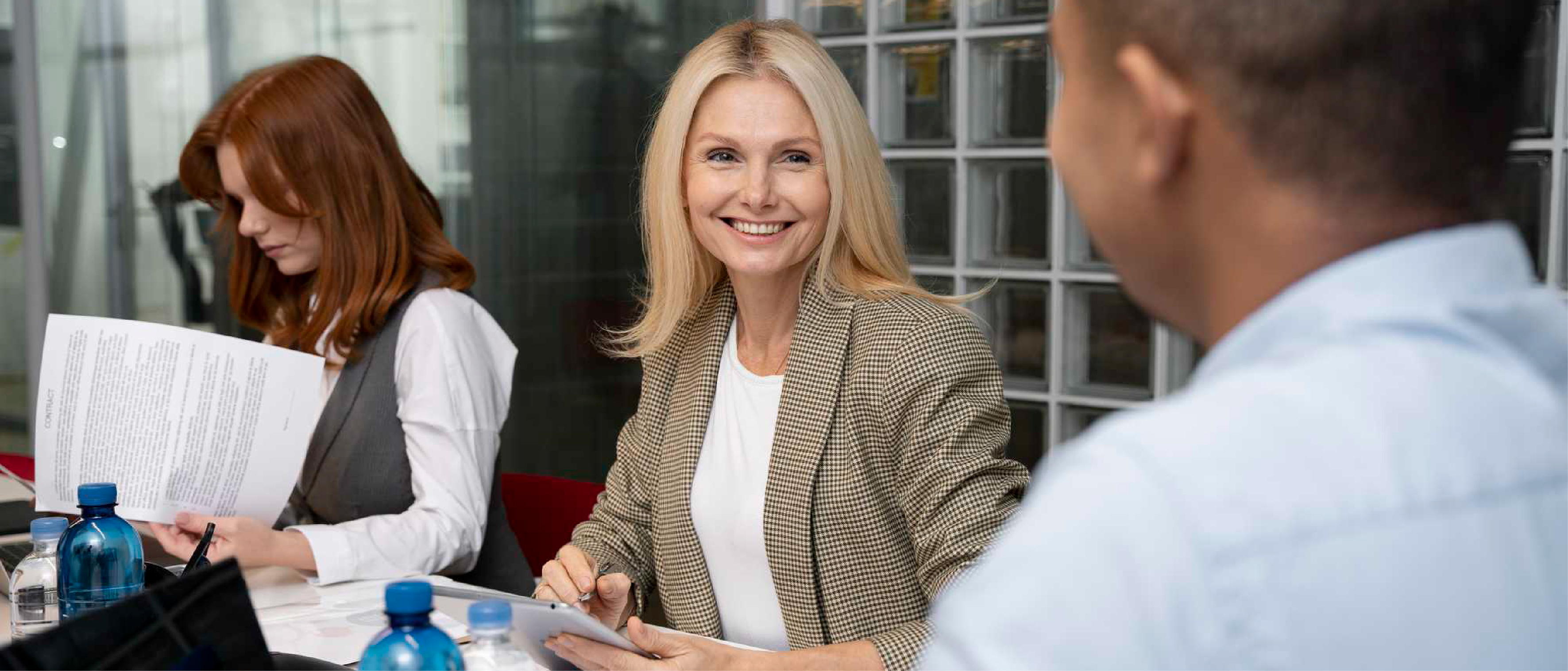 Trzy osoby podczas spotkania w biurze. Na pierwszym planie uśmiechnięta blondynka w beżowej marynarce rozmawia z mężczyzną, obok nich kobieta przegląda dokumenty