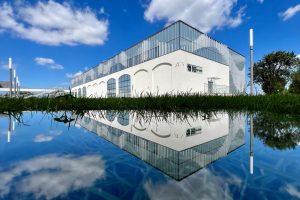 Nowoczesny biały budynek o geometrycznej bryle z przeszklonym piętrem, odbijający się w tafli wody na pierwszym planie. Na jasnej elewacji widoczne są ozdobne łuki oraz logotyp Muzeum Początków Państw