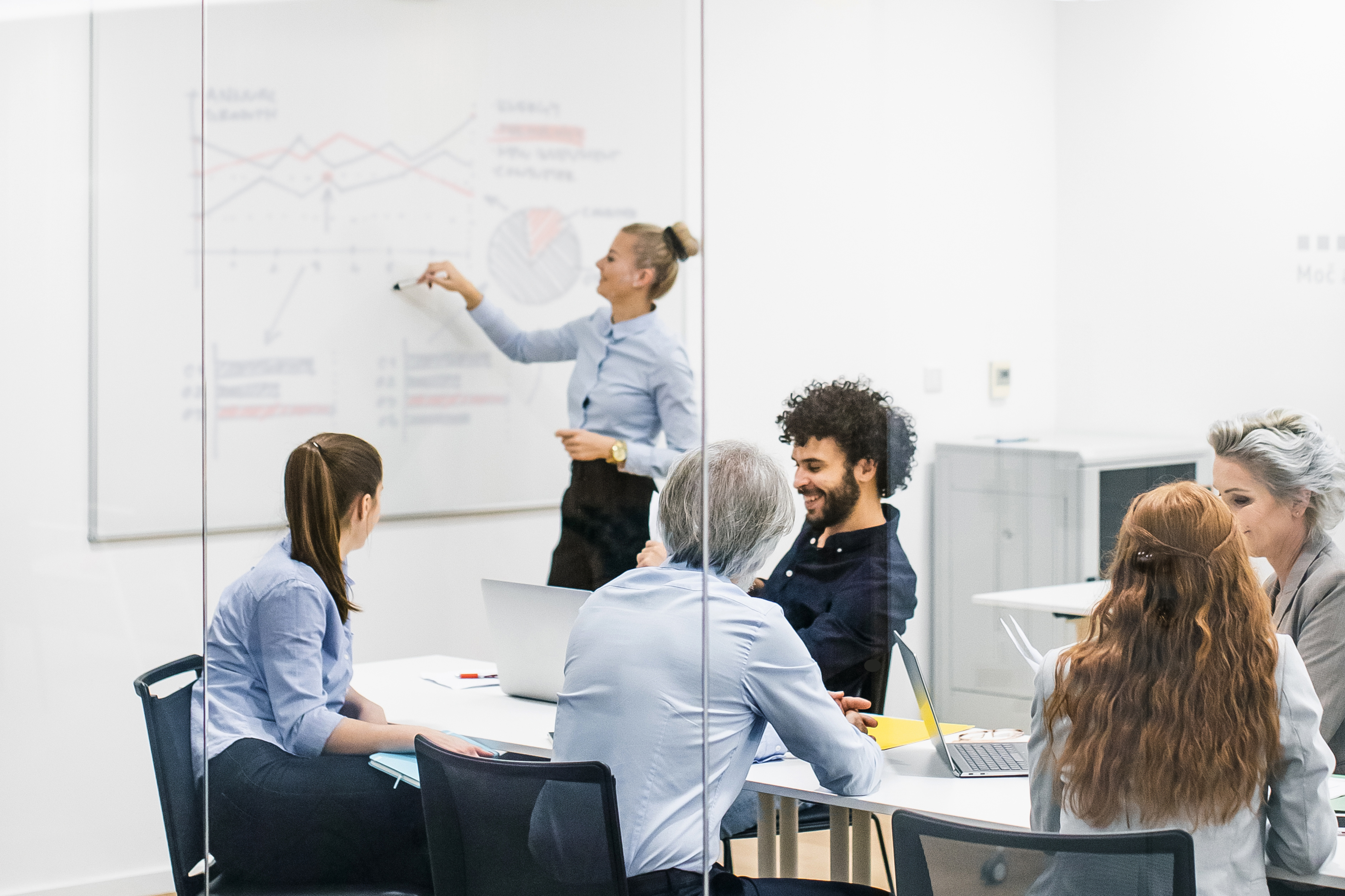 Zespół w firmie siedzący przy stole konferencyjnym słuchający kobiety prowadzącej spotkanie, która stoi przy tablicy