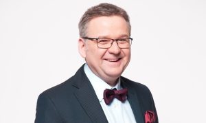 Uśmiechnięty mężczyzna w okularach, ubrany w ciemną marynarkę, jasną koszulę oraz bordową muchę z dopasowaną poszetką, pozujący na białym tle.