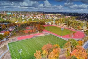 Widok z lotu ptaka na kompleks sportowy - dwa zielone boiska piłkarskie, bieżnię lekkoatletyczną oraz korty tenisowe otoczone drzewa w jesiennych kolorach