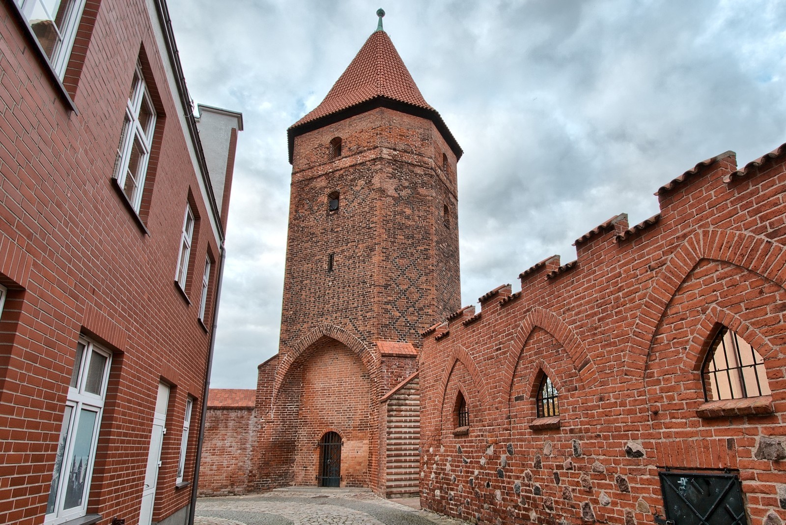 Zdjęcie przedstawia zabytkową Basztę Bluszczową w Lęborku – gotycką, ceglaną wieżę z XIV wieku z dachem namiotowym. Obok widoczny jest zrekonstruowany fragment murów obronnych z blankami i ostrołukowy