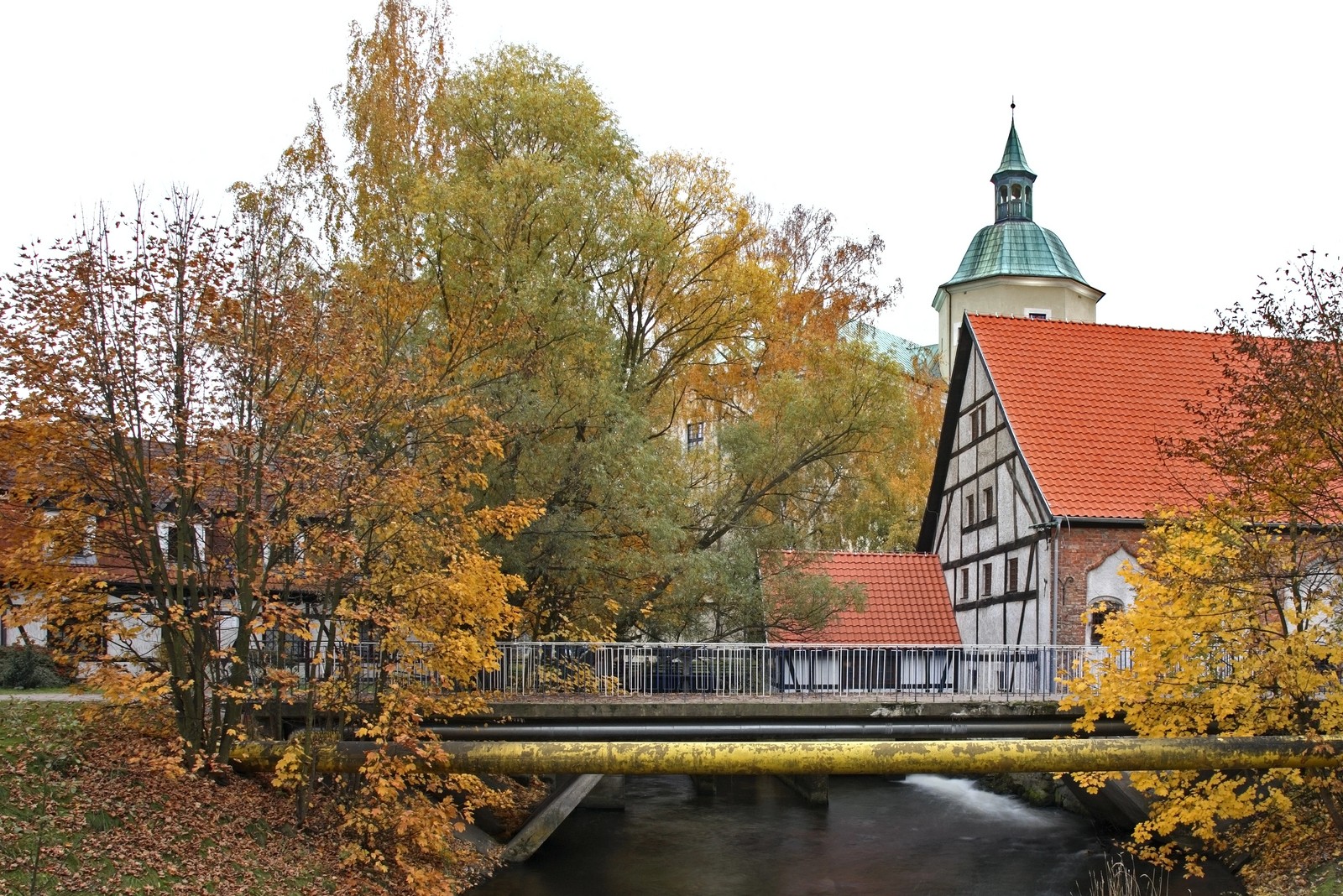 Jesienny widok na rzekę Słupię w Słupsku. Na pierwszym planie widać ciemną wodę płynącą pod mostkiem oraz charakterystyczną, grubą żółtą rurę biegnącą równolegle do kładki.