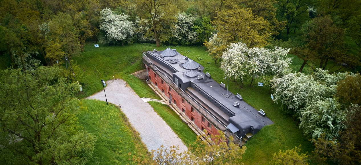 Widok z lotu ptaka na zabytkowy, ceglany fort częściowo ukryty wśród gęstych, zielonych drzew