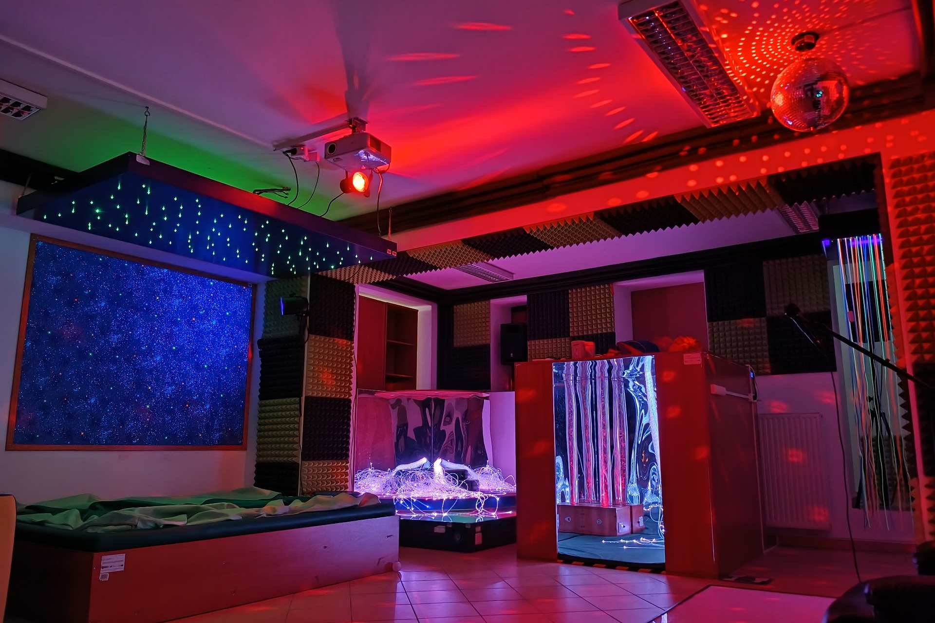 Sala sensoryczna z kolorowym oświetleniem, projektorem świetlnym i elementami do terapii oraz relaksu, takimi jak podświetlane panele, materace i światłowody sensoryczne.