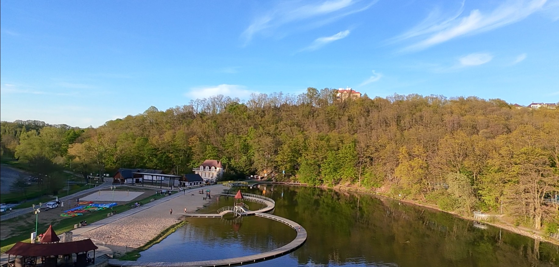 Panoramiczny widok na spokojny zalew otoczony lasem. Na pierwszym planie znajduje się piaszczysta plaża i drewniane pomosty o falistym kształcie prowadzące nad wodą. W tle widać wzgórze porośnięte drz