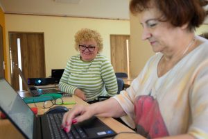 Podczas zajęć w Klubie Rozwoju Cyfrowego w Swarzędzu uczestniczki pracują przy laptopach. Jedna z nich pisze na klawiaturze, a druga z uśmiechem obserwuje jej działania.
