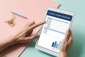 Grafika z ludzkimi dłońmi trzymającymi tablet z ankieta z napisem Interreg Europa Środkowa